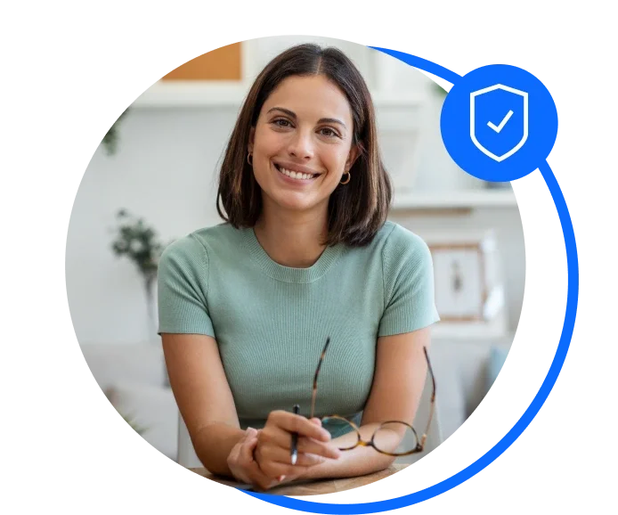 Brunette female sat down working at a desk at home. She's holding her glasses, wearing a pale green top and smiling to the camera.